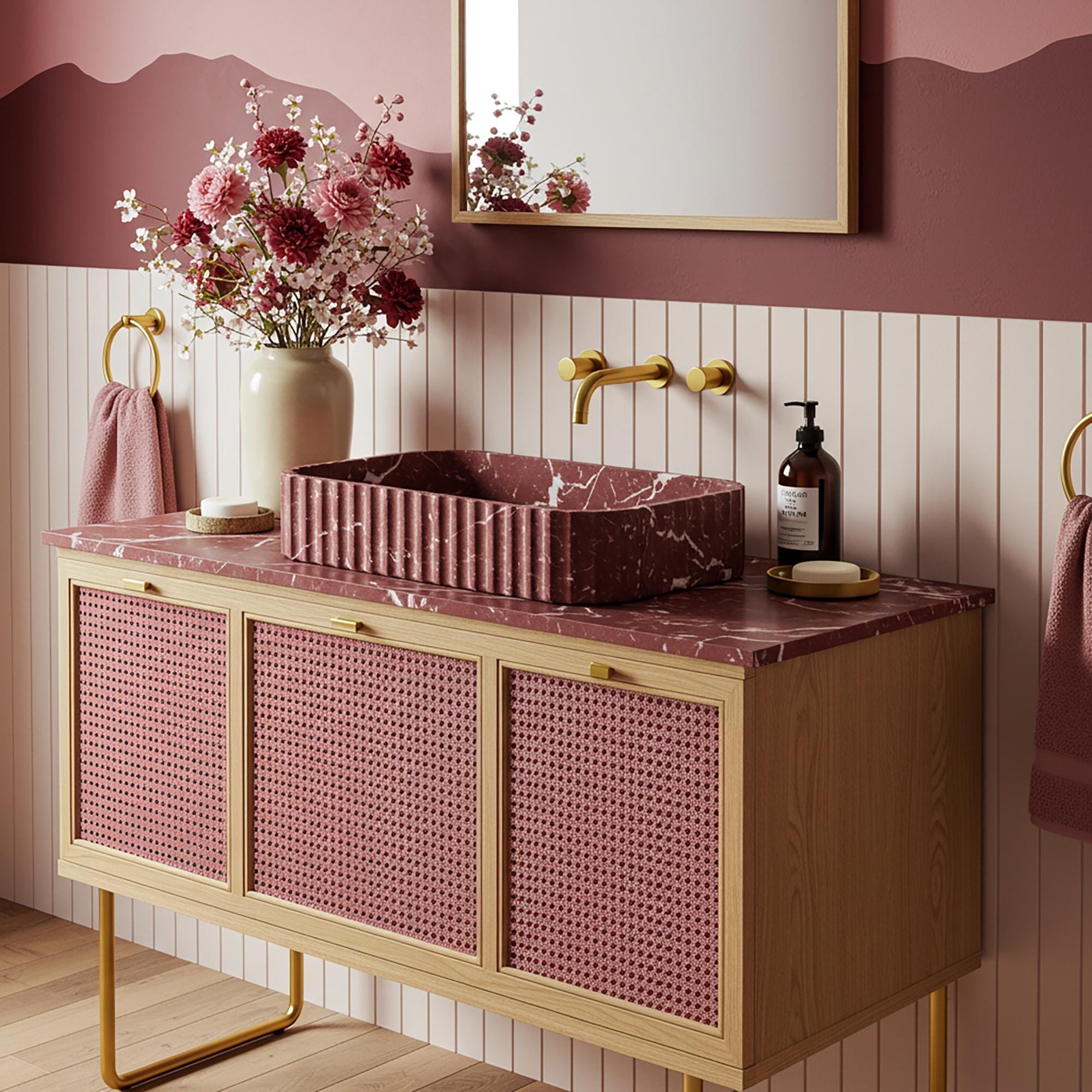 A bohemian chic red marble vanity with a fluted stone sink, set on a wooden cabinet with rattan doors and brass fixtures in a warm, softly styled bathroom.
