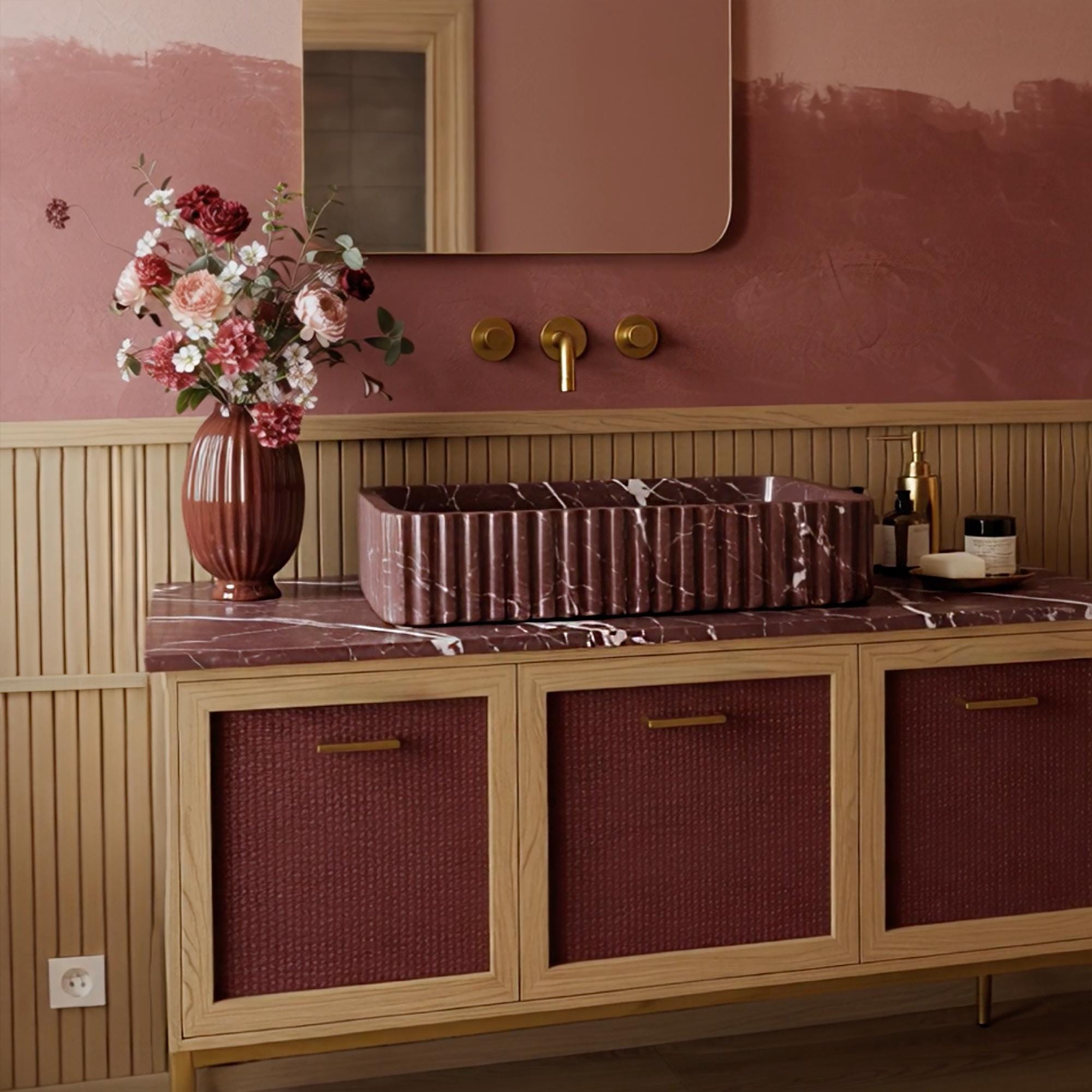 A sculptural Rosso Levanto marble vanity featuring a fluted stone basin and a burgundy rattan cabinet in a timeless bathroom design.
