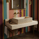 A fluted beige travertine console sink with vertical carving, set on a brass stand against a colorful striped wall with warm brass fixtures.
