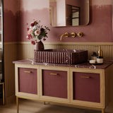 A fluted Rosso Levanto marble vanity with deep burgundy veining, paired with a rattan-front cabinet and brass accents in a warm, refined bathroom interior.
