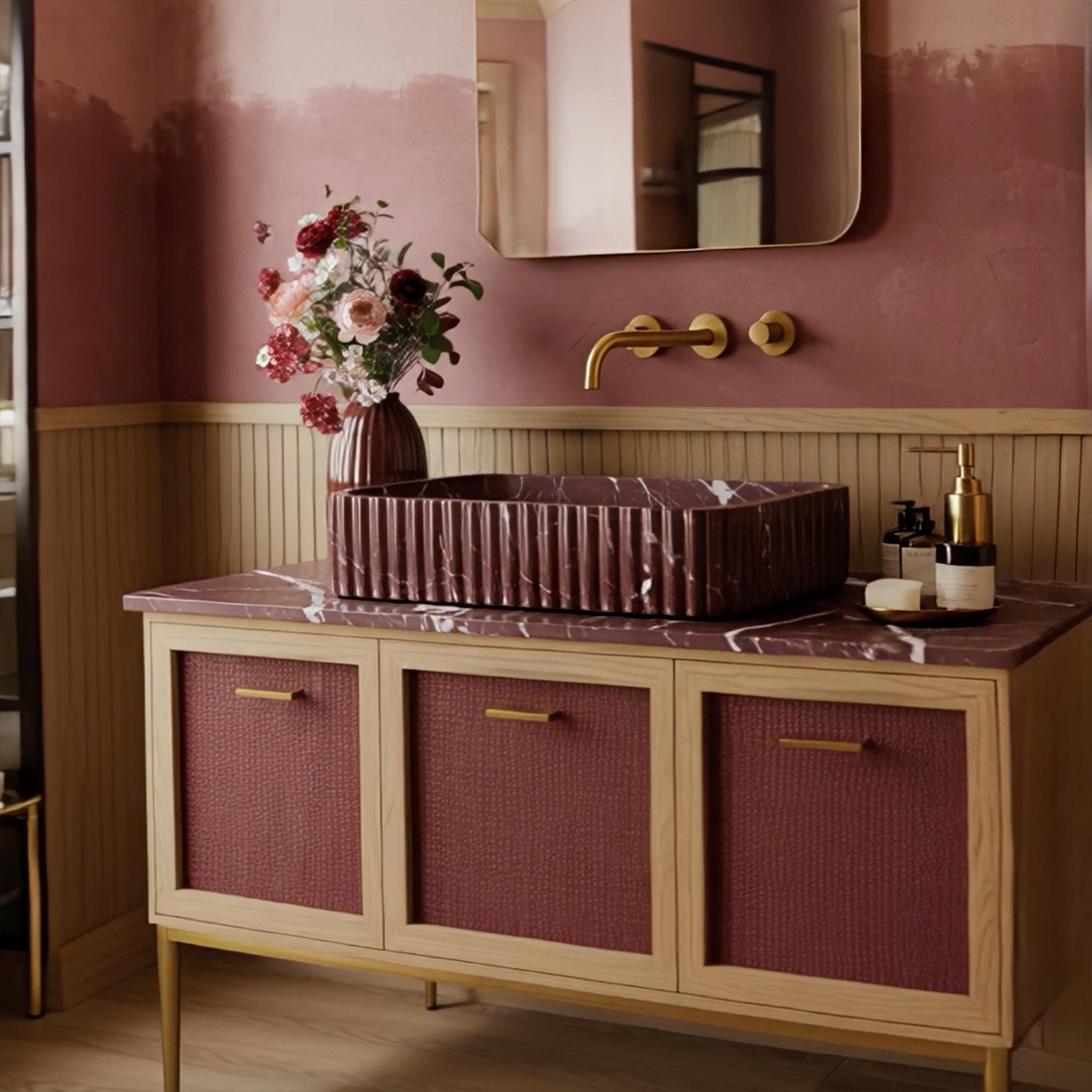 A fluted Rosso Levanto marble vanity with deep burgundy veining, paired with a rattan-front cabinet and brass accents in a warm, refined bathroom interior.

