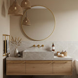 A Tundra Grey marble floating vanity with a stepped block sink, featuring subtle white veining and oak wood drawers with brass hardware in a calm bathroom setting.
