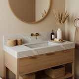 A Tundra Grey marble integrated sink vanity with soft linear veining, paired with an oak wood console and brass wall-mounted faucet in a calm bathroom setting.
